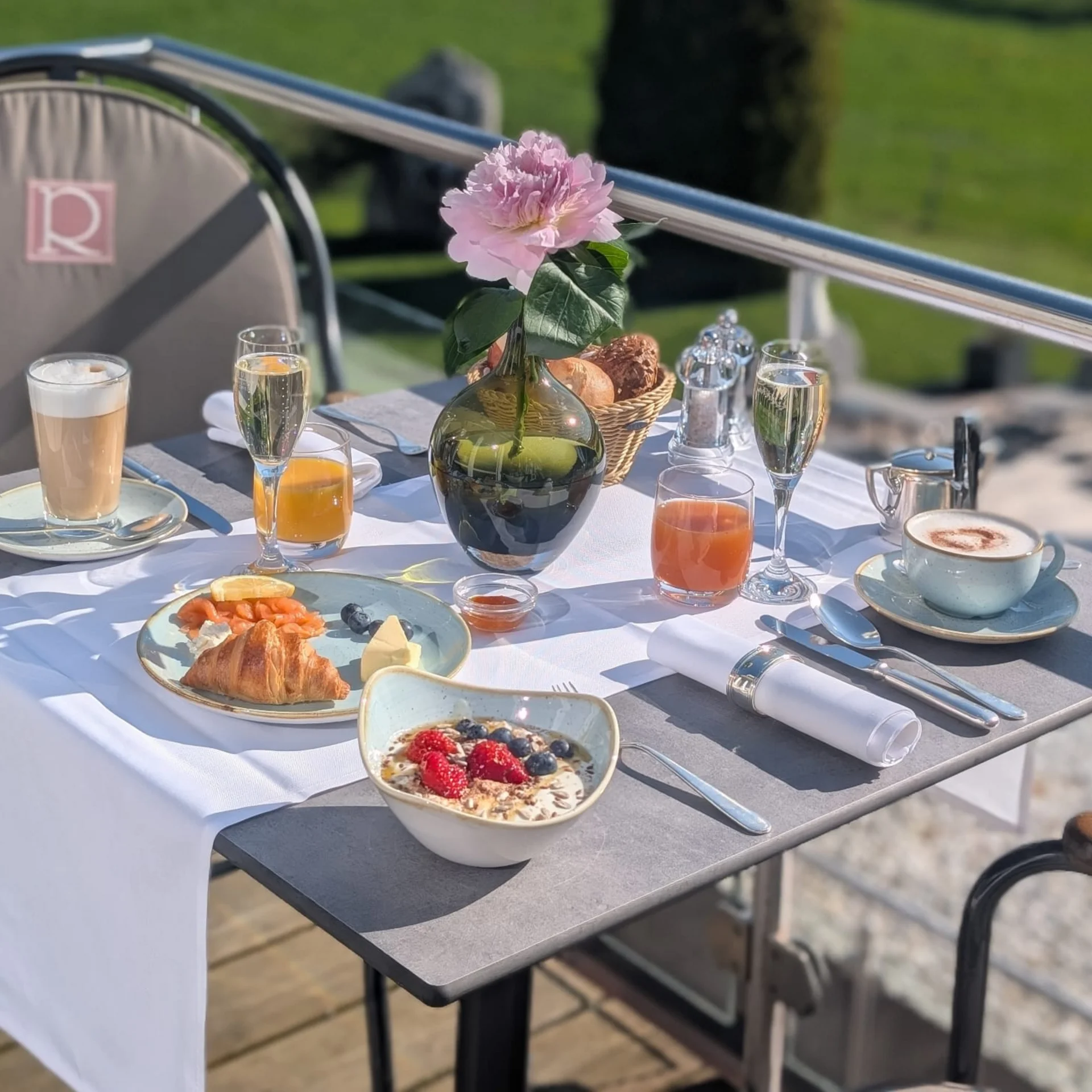 Tisch mit Frühstück und rosa Blume auf Terrasse mit grünem Hintergrund