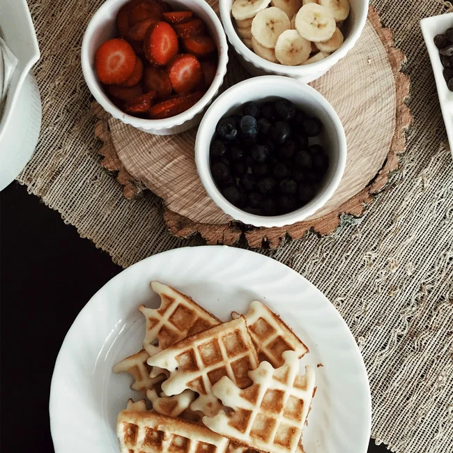 Waffles on white plate with bowls of strawberries, bananas, and blueberries