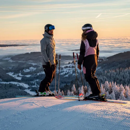 Two skiers watch the sunrise over a snow-covered mountain landscape