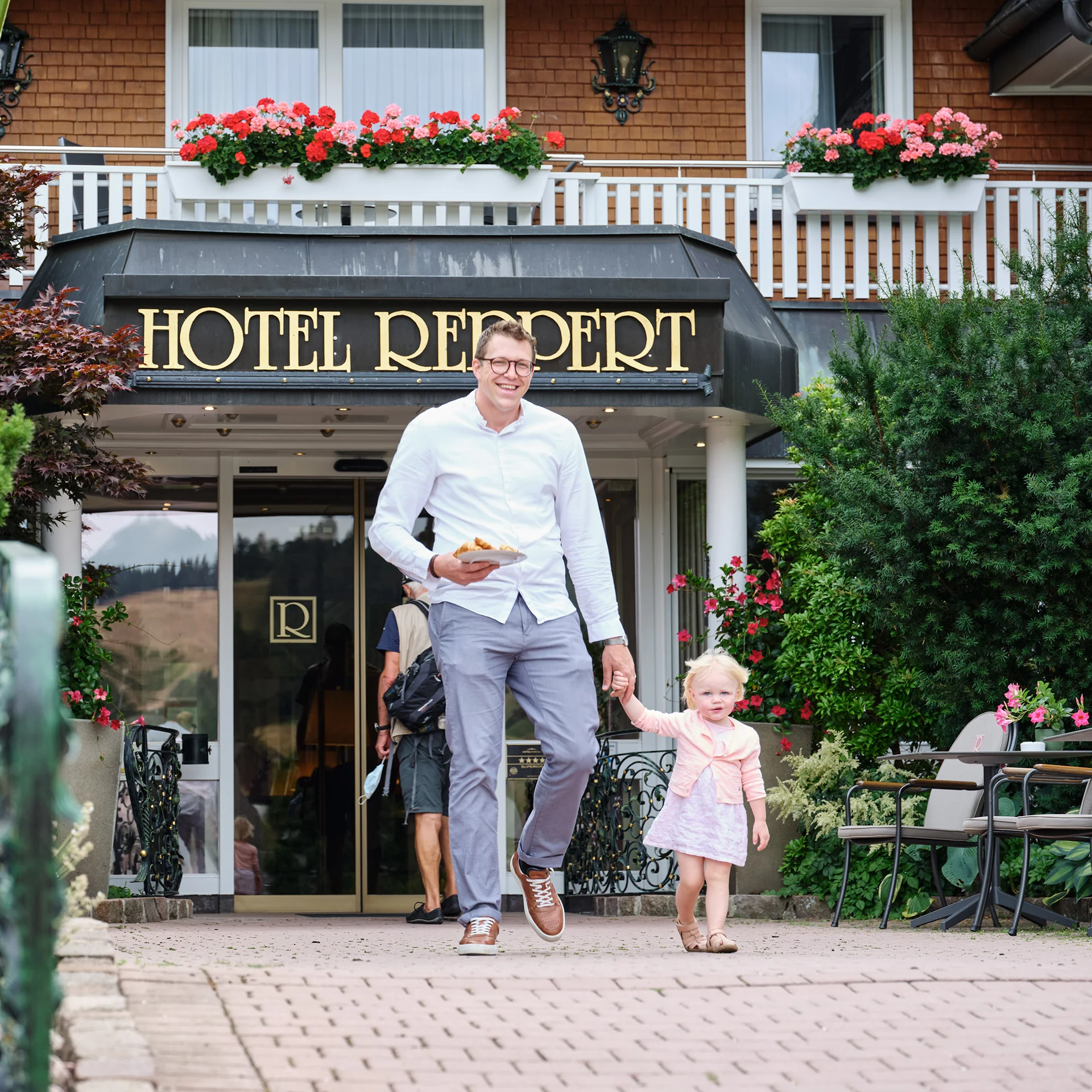 Mann mit Kind vor Hotel Reipert, umgeben von Blumen und Grünpflanzen