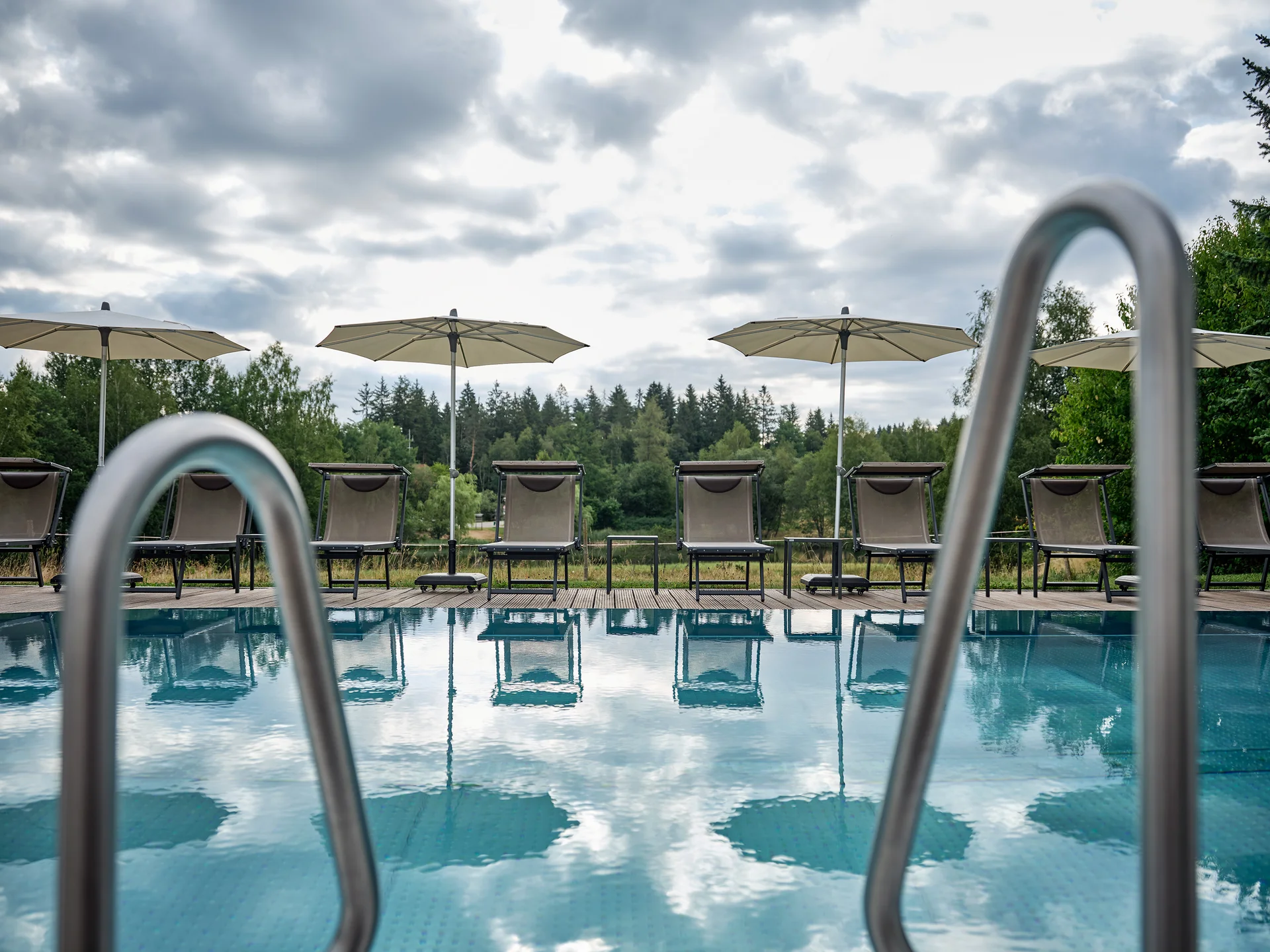 Leerer Pool mit Liegestühlen und Sonnenschirmen vor bewölktem Himmel und Bäumen