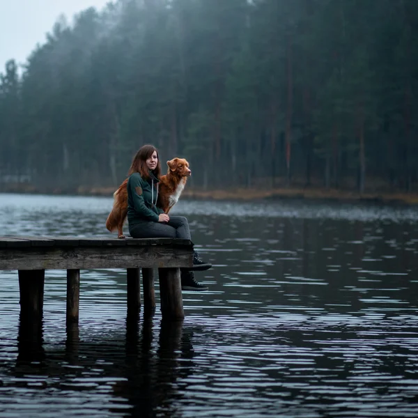 Frau sitzt mit Hund auf Steg am See vor einem bewaldeten Ufer
