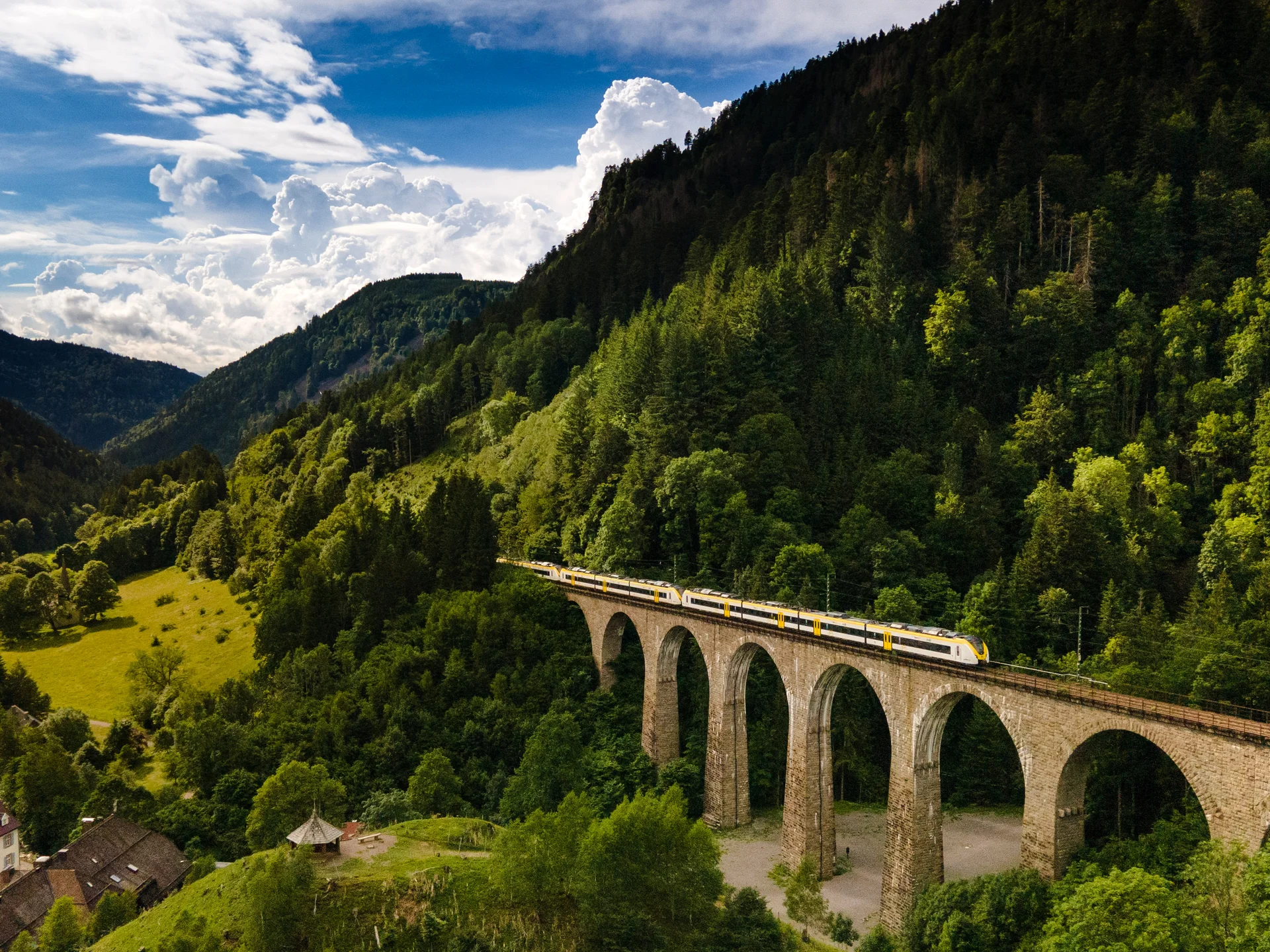 Zug fährt über einen Steinbogenviadukt in bewaldeten Bergen bei klarem Himmel