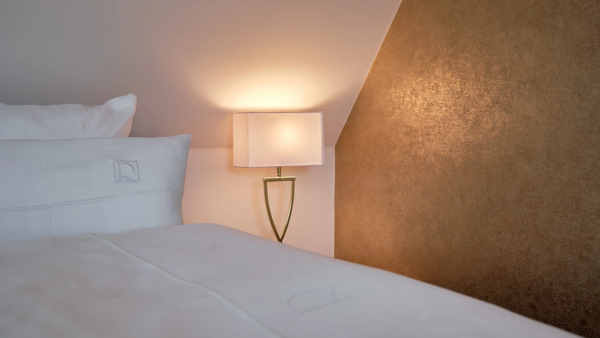 Weißes Bett mit Kissen neben einer leuchtenden Nachttischlampe und goldenem Wandpaneel