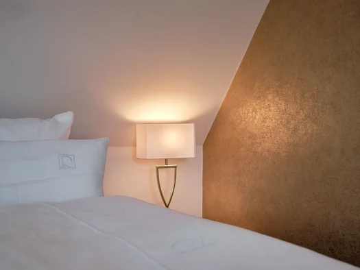 Weißes Bett mit Kissen neben einer leuchtenden Nachttischlampe und goldenem Wandpaneel