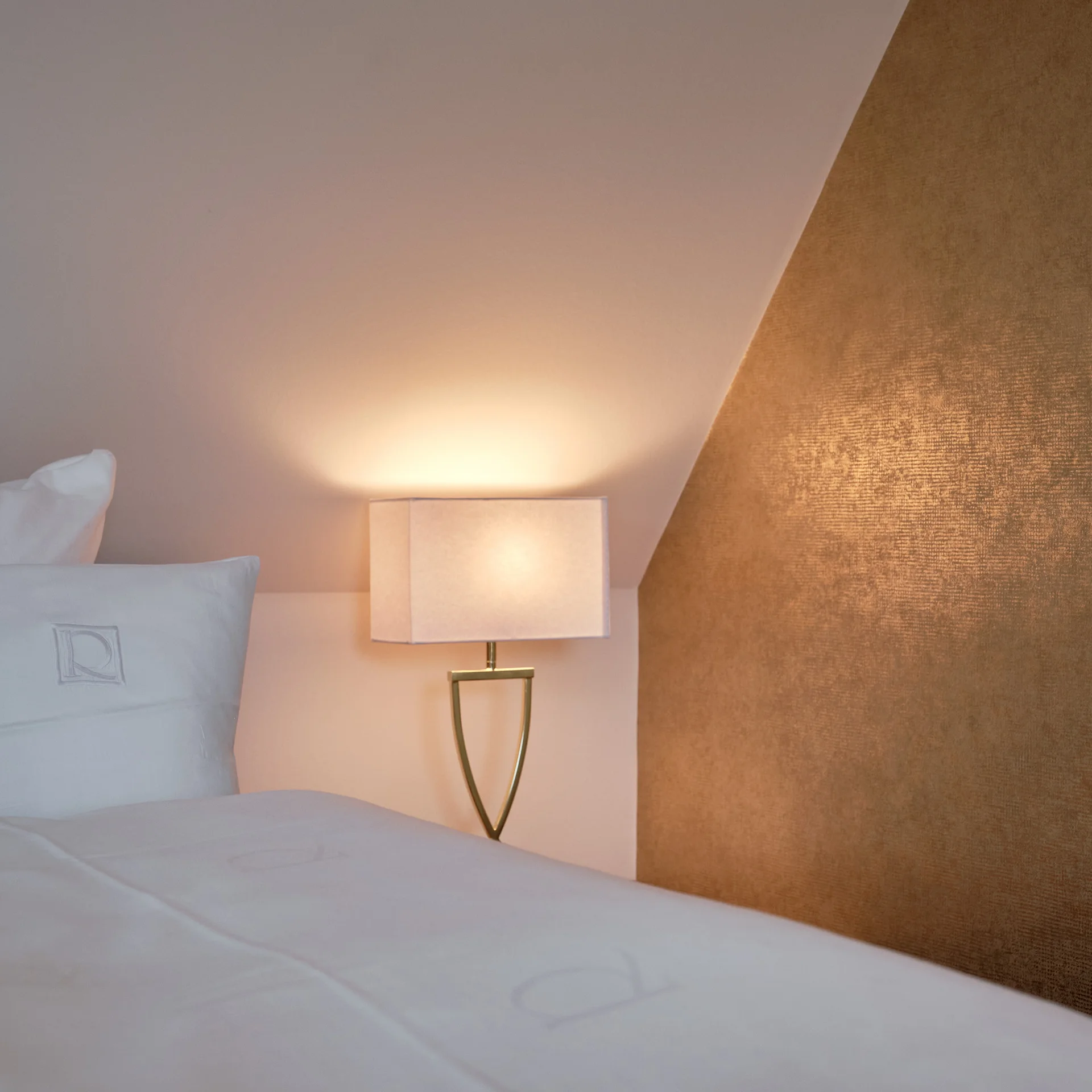 Weißes Bett mit Kissen neben einer leuchtenden Nachttischlampe und goldenem Wandpaneel