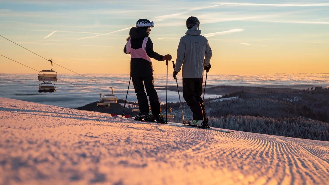 Zwei Skifahrer auf einer verschneiten Piste mit Bergblick im Abendlicht