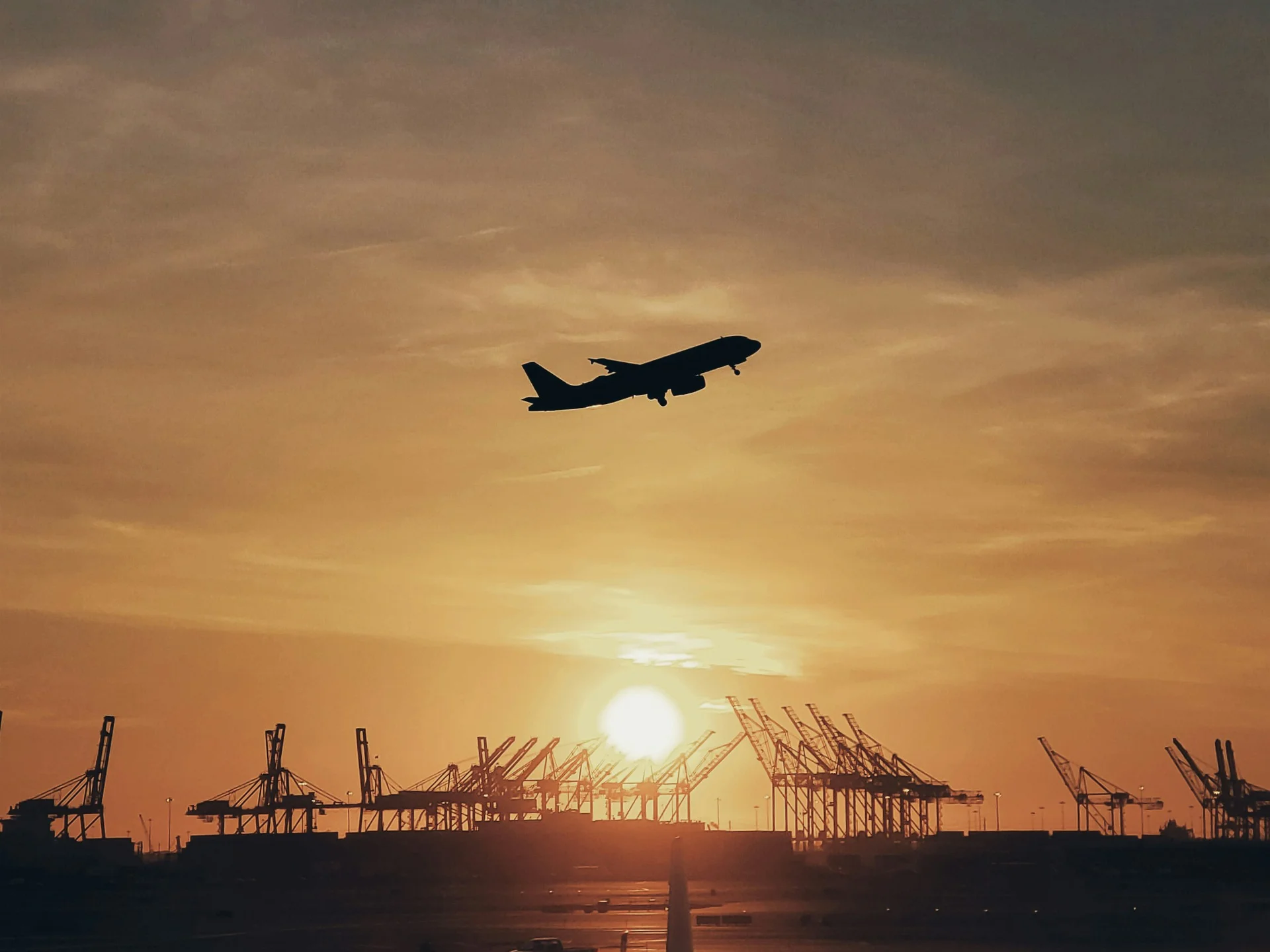 Flugzeug startet bei Sonnenuntergang über einem Hafen mit Kränen