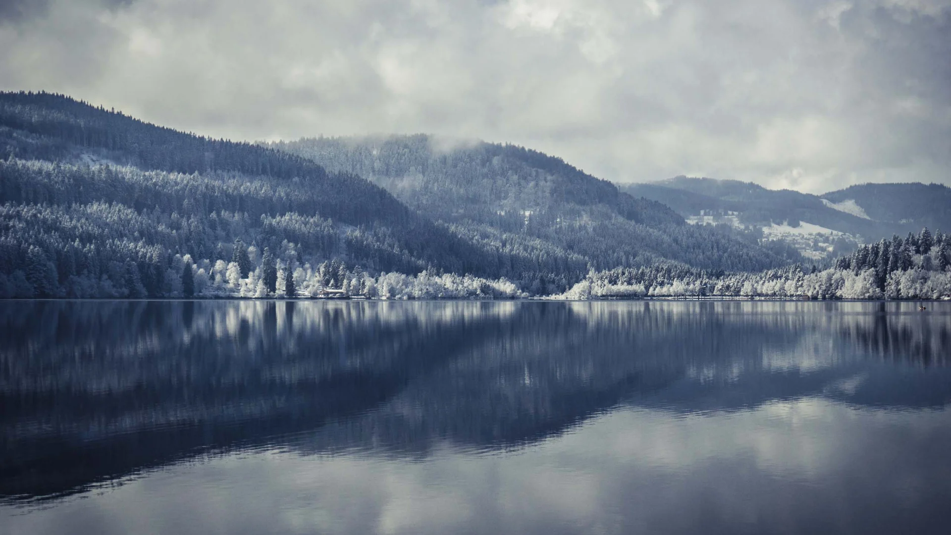 Verschneite bewaldete Hügel spiegeln sich in einem ruhigen See unter bewölktem Himmel