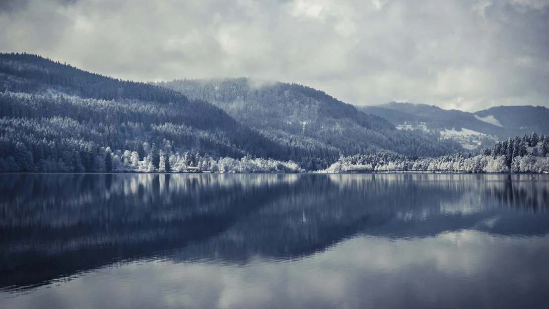 Des collines boisées enneigées se reflètent dans un lac calme sous un ciel nuageux