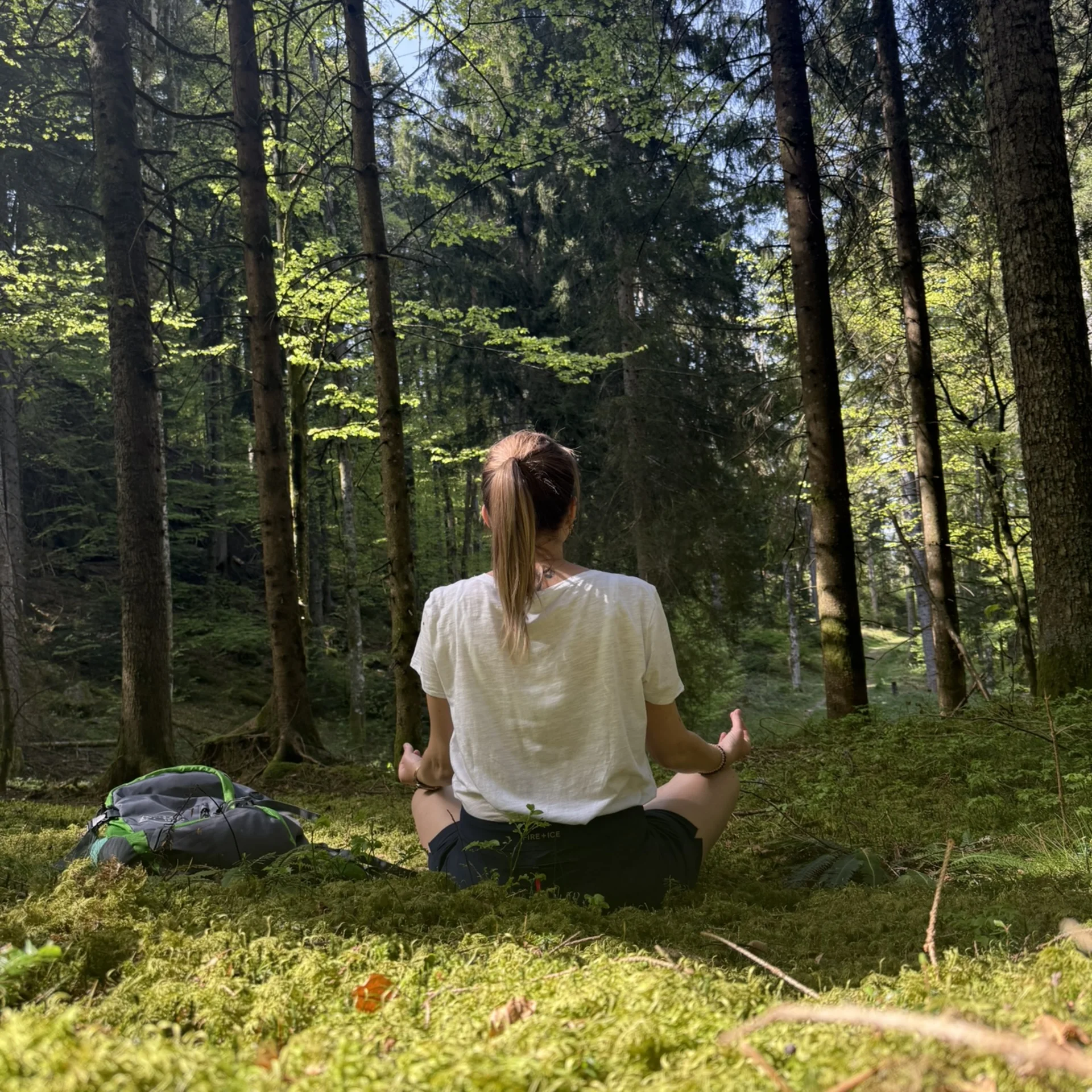 Person meditiert im grünen Wald, sitzend auf Moos mit Rucksack neben ihr