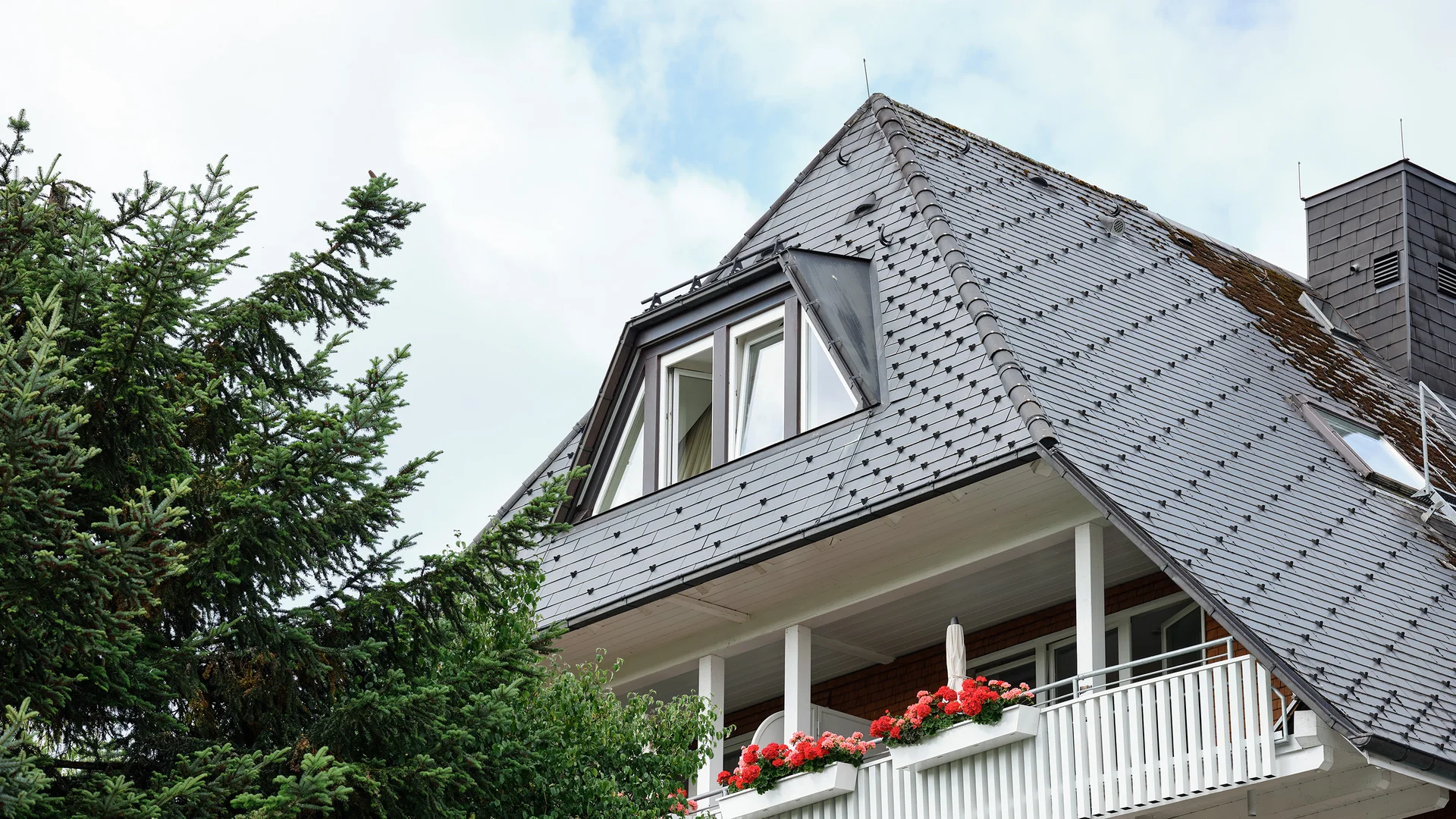 Haus mit schiefergedecktem Dach und blühenden Blumen auf Balkonen