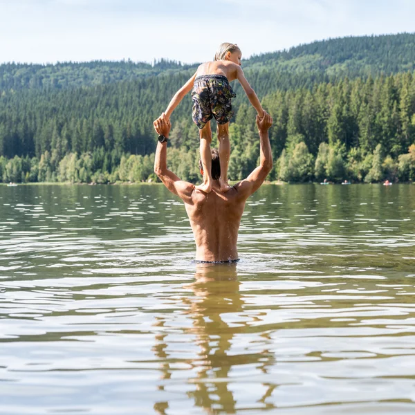 Mann hebt Kind beim Schwimmen in einem See vor bewaldeten Bergen