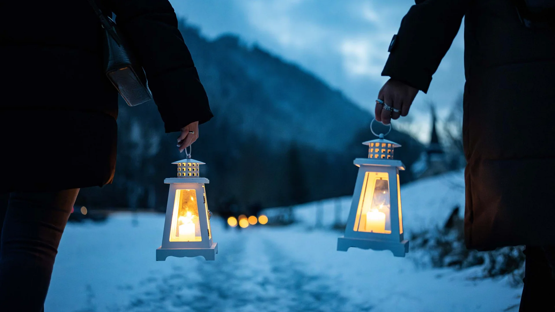 Deux personnes portent des lanternes allumées sur un chemin enneigé, devant des montagnes