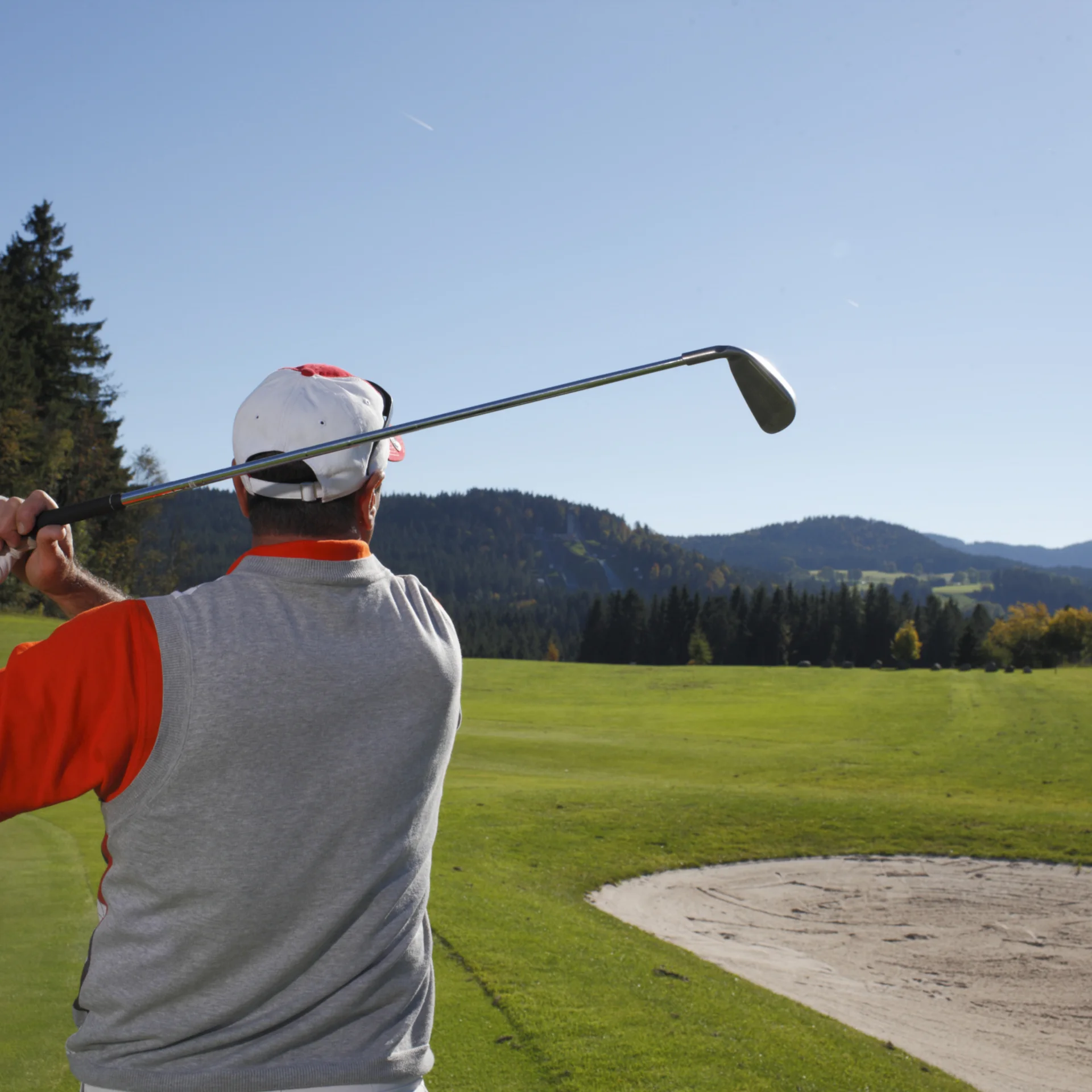 Golfspieler von hinten beim Abschlag auf einem grünen Golfplatz mit Bergen