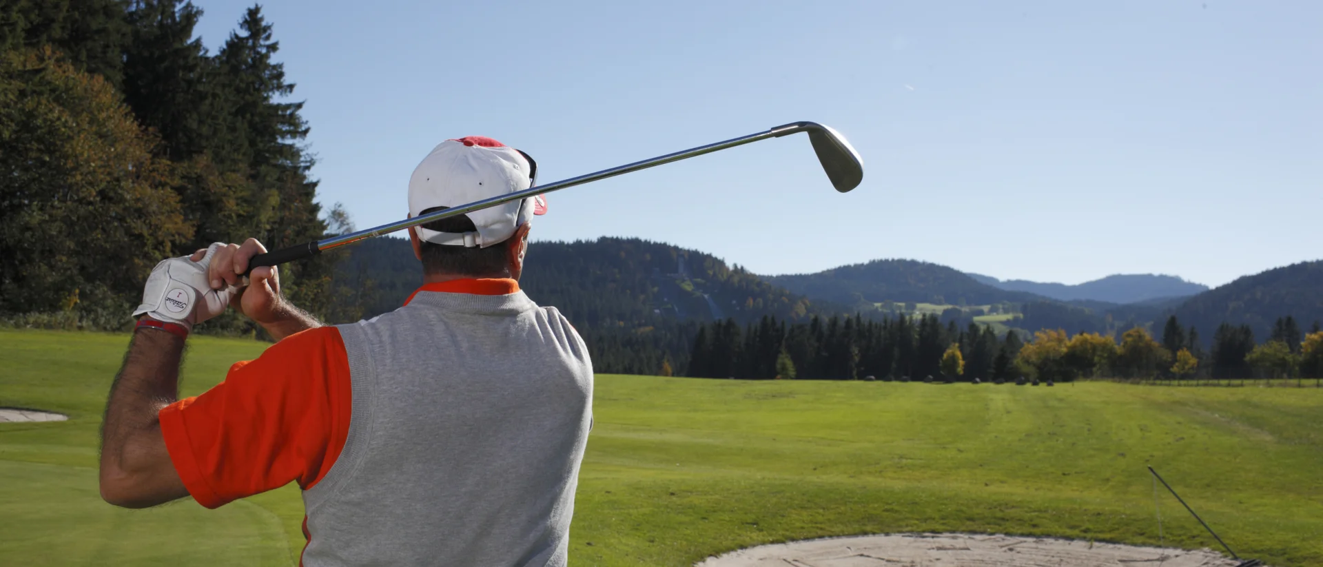 Golfspieler von hinten beim Abschlag auf einem grünen Golfplatz mit Bergen