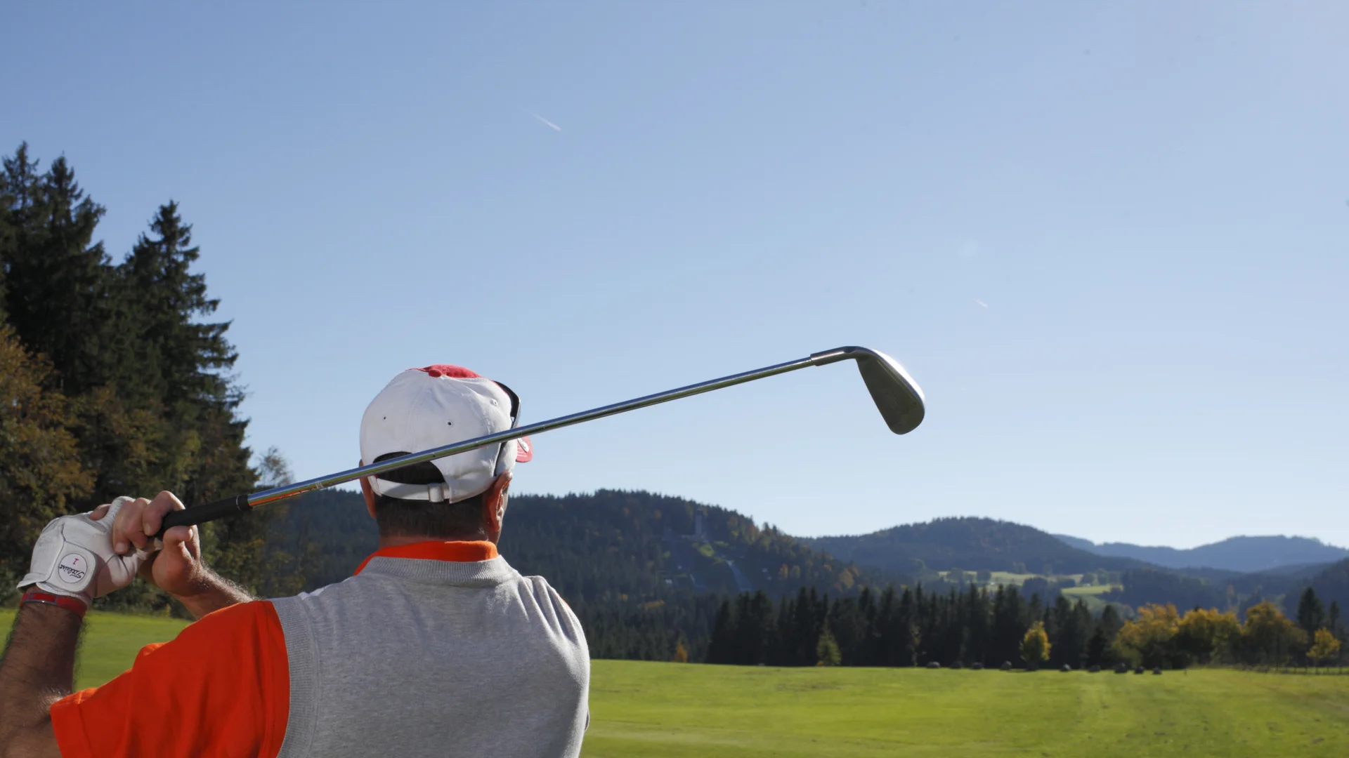 Golfspieler von hinten beim Abschlag auf einem grünen Golfplatz mit Bergen