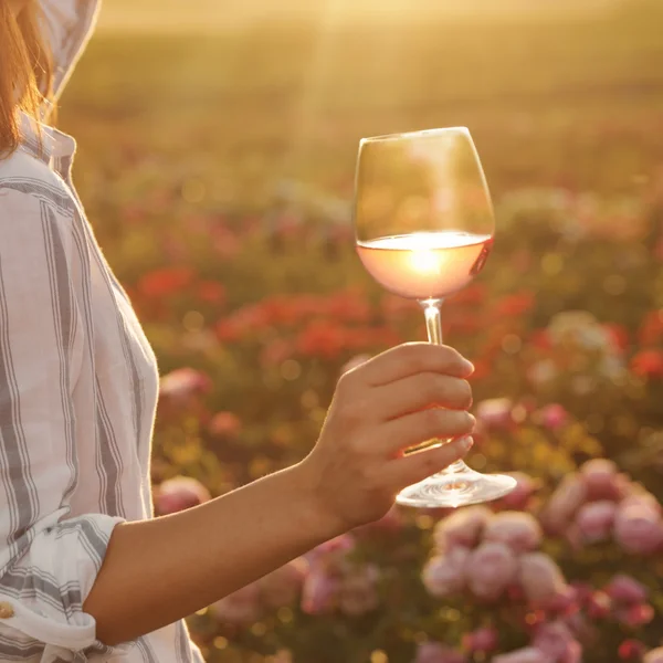Frau hält ein Weinglas mit Roséwein im Sonnenlicht über einer blühenden Wiese