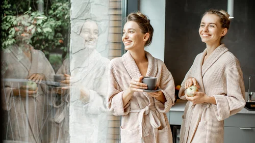 Zwei lächelnde Frauen im Bademantel mit Tasse und Apfel am Fenster